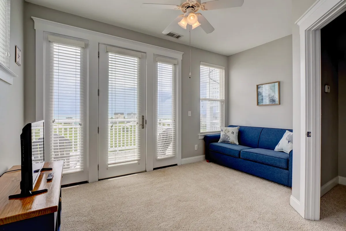 A relaxing living area with a plush blue sofa, ample natural light streaming in through expansive windows, a ceiling fan, and a small table. The space looks perfect for quiet mornings or gathering with friends while enjoying views beyond the glass.