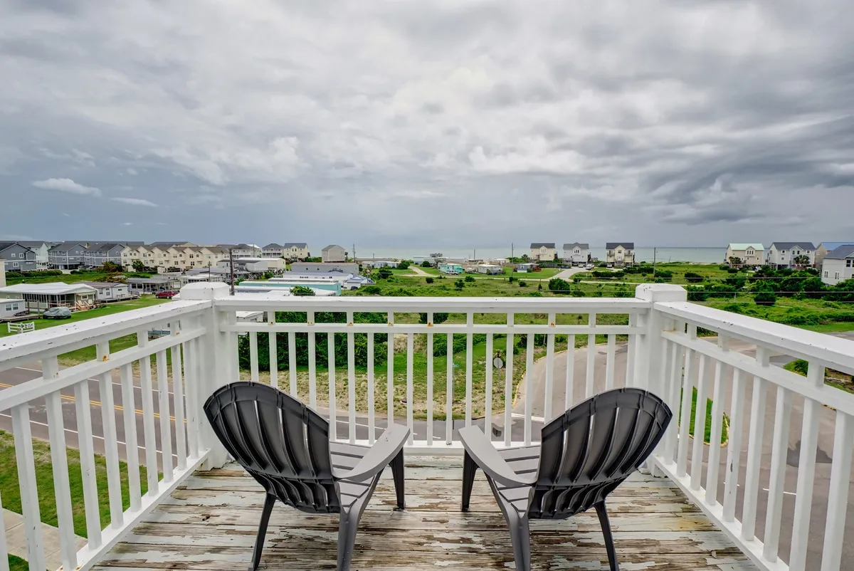 The image captures two inviting chairs on a private balcony overlooking lush greenery and rooftops, with the tranquil blue ocean stretching out in the distance. The scene suggests a peaceful place to relax, enjoy the fresh sea breeze, and take in sweeping coastal views.