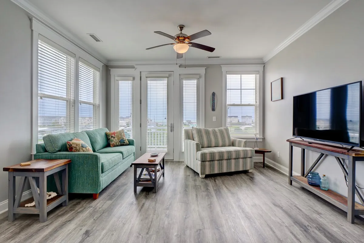 A comfortable living area with a sofa, armchair, and coffee table oriented toward large windows or a television. The room is bathed in natural light, suggesting a seamless connection between indoor relaxation and the outdoor decks beyond.