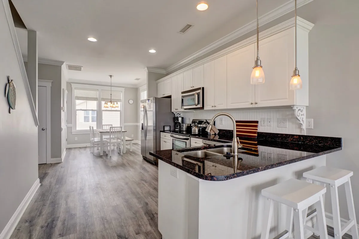 A bright, modern kitchen is seen here with a large island at the center, flanked by white chairs and stainless-steel appliances. The dining area, visible just beyond the kitchen, promises plenty of room for shared meals and memorable moments, with chic light fixtures contributing to a fresh, updated ambiance.