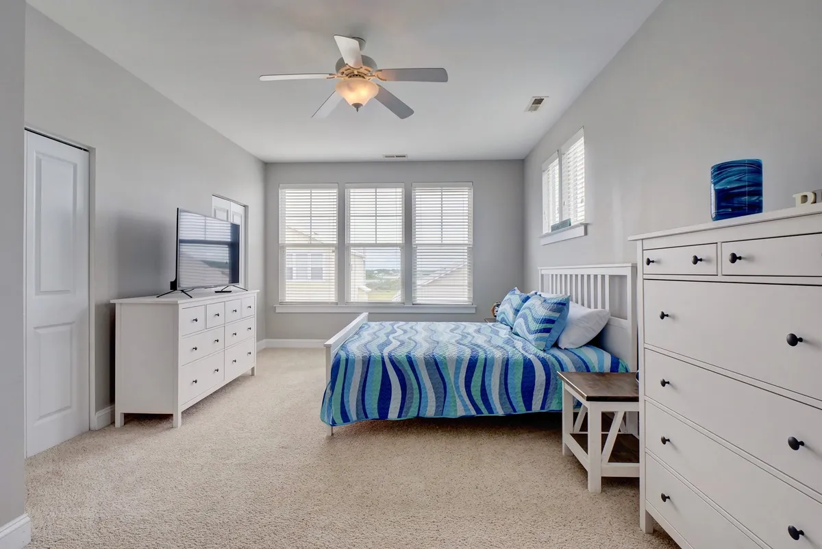 A serene bedroom with a bed dressed in blue striped bedding, large windows with blinds allowing natural light, a white dresser, and a TV on a stand. The environment is peaceful and tailored for quiet rest.