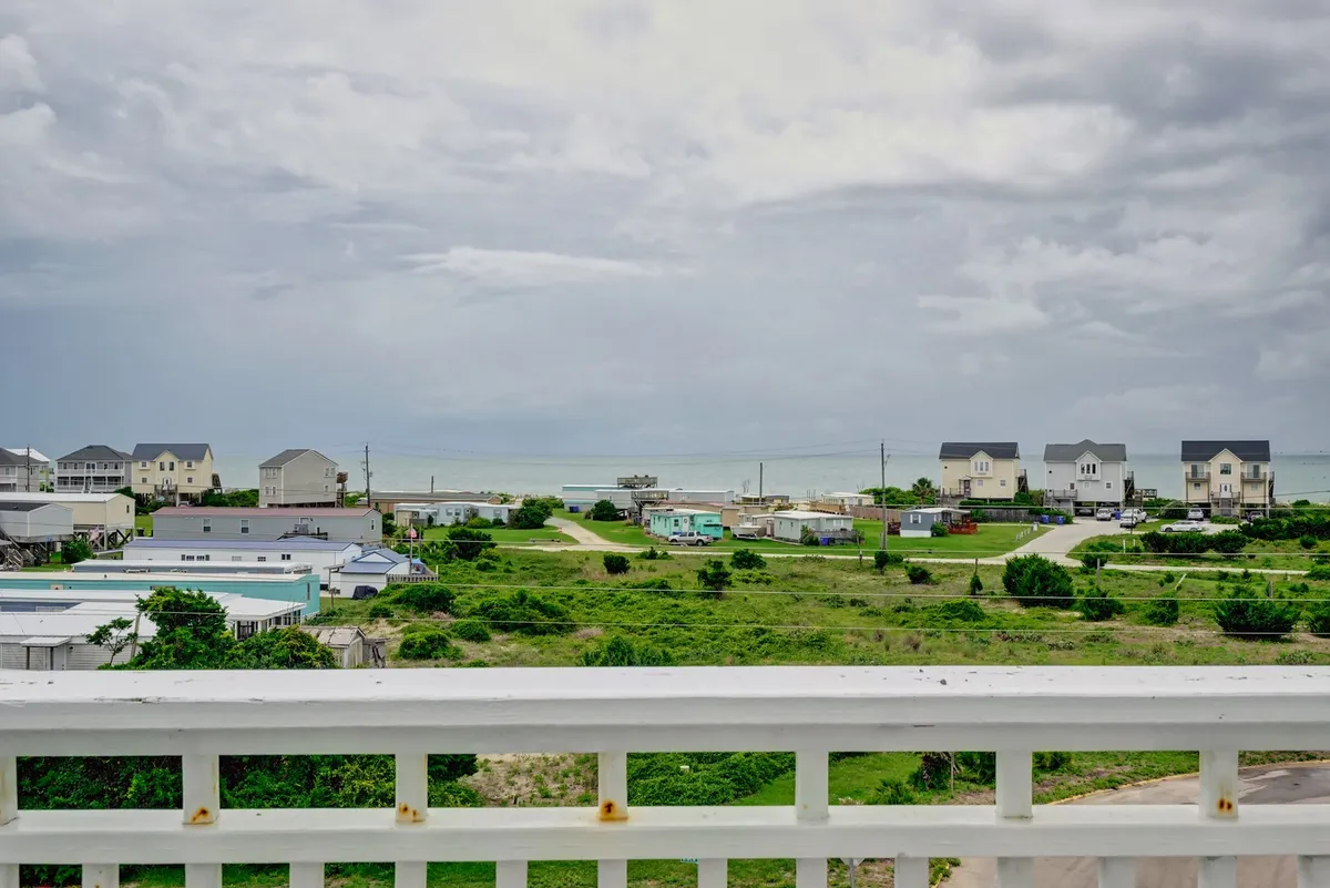 A view from a deck or balcony highlighting a sweeping ocean vista with distant houses and lush greenery under a tranquil sky. The scene evokes the ambiance of sitting outdoors, gazing at the ocean, and enjoying moments of relaxation, sunrise coffees, or sunset drinks.