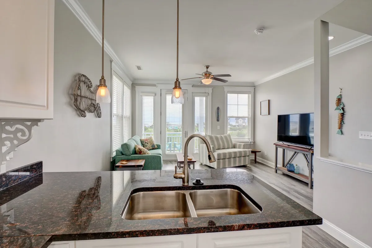 The open-concept space seamlessly blends a modern kitchen with a cozy living area, featuring a green sofa, striped armchair, and a television—creating the perfect setting for relaxation after meals or group activities. Hanging pendant lights and generous windows maintain a bright, lively atmosphere.